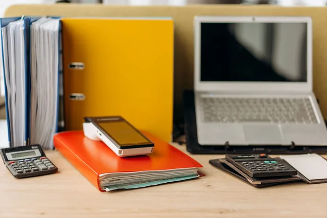 Mesa de trabalho com computador, calculadoras e pastas, representando uma operação administrativa mais estruturada
